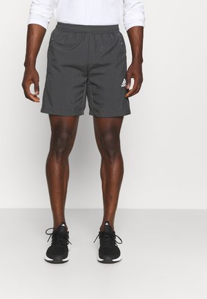 Mann in schwarzen Adidas Sportshorts und schwarzen Laufschuhen, der vor einem schlichten weißen Hintergrund steht.