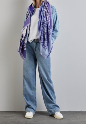 Femme portant un jean large bleu clair, des baskets blanches, un haut blanc, une veste en jean et une grande écharpe violette à motifs, debout contre un mur gris.