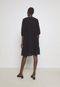 Robe noire mi-longue ornée d'un motif broderie anglaise, avec des manches trois-quarts et un ourlet à volants. Associée à des sandales à talons bloqués et dos ouvert.