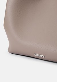 Geantă texturată bej cu un finisaj neted, având un logo argintiu "DKNY" în față. Formă curbată și detalii de design minime.