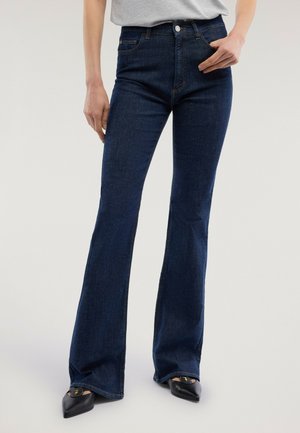 Frau trägt dunkelblaue hochtaillierte Schlagjeans, graues eingestecktes Oberteil und schwarze spitze Ballerinas, steht vor neutralem Hintergrund.