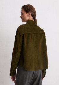 Chemise en velours côtelé vert olive avec col, présentant une coupe ample, une texture côtelée verticale et des poignets à boutons. Détail de l'emmanchure visible.