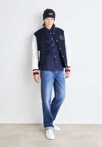 Giacca varsity blu navy e bianca con logo, abbinata a una maglietta scura e jeans blu, completata da sneakers bianche e un berretto blu navy.