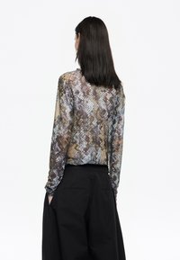 Langearmige, doorzichtige blouse met een textuur en een multicultureel geometrisch patroon. Gecombineerd met hoge zwarte broeken, waarbij de achterkant zichtbaar is.