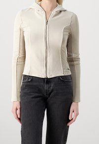 Kvinna iklädd beige zip-up hoodie med ribbade sidopaneler och svarta högmidjade jeans, stående mot en enfärgad bakgrund.