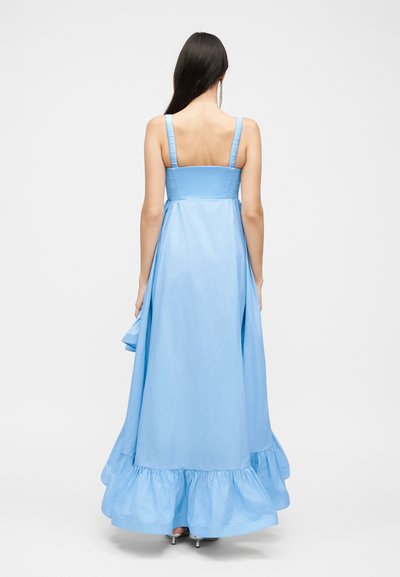 Γυναίκα με μακριά σκούρα μαλλιά που φοράει ένα ανοιχτό μπλε αμάνικο μακρύ φόρεμα με βολάν στη βάση, στέκεται με την πλάτη σε ένα μονόχρωμο φόντο.