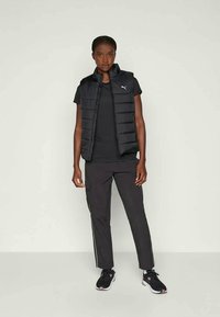 Gilet puffa nero con colletto alto, indossato sopra una maglietta nera e abbinato a pantaloni cargo scuri. L'outfit include scarpe da ginnastica nere.