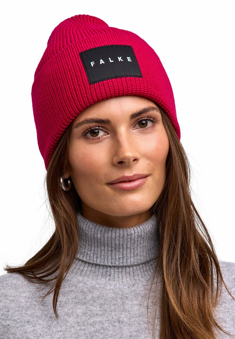 Donna dai lunghi capelli castani indossa un orecchino a cerchio in argento, un maglione a collo alto grigio a coste e un berretto di maglia rosa acceso con etichetta nera "FALKE".