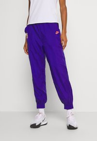 Fialové běžecké kalhoty Nike z lehkého materiálu, s elastickým pasem a manžetami, s oranžovým logem a detaily bočních kapes.