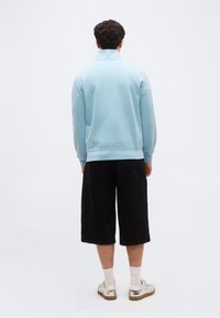 Άνδρας που στέκεται γυρισμένος με την πλάτη, φοράει γαλάζιο πουλόβερ, μαύρες βερμούδες μέχρι το γόνατο, λευκές κάλτσες και λευκά αθλητικά παπούτσια με καφέ σόλες.