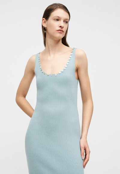 Νεαρή γυναίκα με αμάνικο ριμπ φόρεμα ανοιχτού μπλε χρώματος με λαμπερά κουμπιά κατά μήκος του λαιμού, φορώντας χρυσούς κρίκους σκουλαρίκια, στέκεται μπροστά σε απλό φόντο.