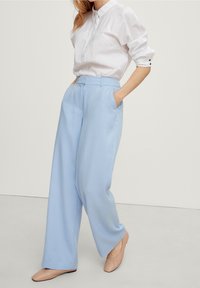 Femme portant une chemise blanche à manches longues, un pantalon large bleu clair et des chaussures plates beiges, se tenant avec les mains dans les poches.