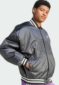 Black bomber jacket made of shiny fabric, featuring a large embossed logo, striped cuffs, and waistband. Limited additional detailing visible.