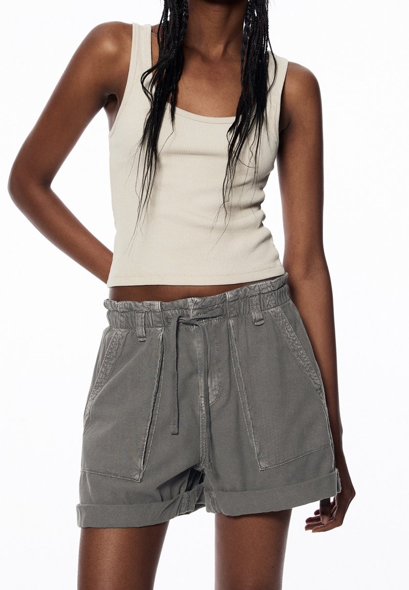 Pantalones cortos grises y ligeros con cintura ajustable, bolsillos laterales y puños remangados. Combinados con una camiseta sin mangas beige ajustada.