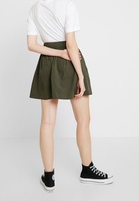 Jupe vert olive avec un taille élastique froncé, longueur genoux, associée à des baskets montantes noires et un t-shirt blanc.