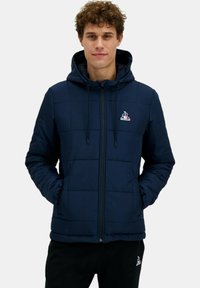 Giacca imbottita navy con cappuccio regolabile, cerniera frontale e tasche laterali. Presenta un piccolo logo sul petto. Texture liscia.