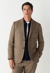 Blazer marron en mélange de laine avec revers crantés, deux poches avant et fermeture à boutons. Porté avec un haut zippé bleu marine en dessous.