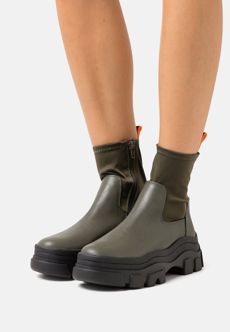 Jambes portant des bottines vert olive à semelles épaisses noires et fermetures éclair latérales, avec des languettes de tirage orange à l'arrière.