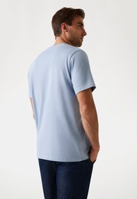Hombre con pelo corto castaño que lleva una camiseta de manga corta azul claro y pantalones oscuros, de pie con las manos en los bolsillos, mirando hacia el lado.