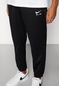 Černé sportovní kalhoty Nike s elastickým pasem, zúženým střihem a bílým logem "AIR" na levém stehně. Materiál je hladký a lehký.