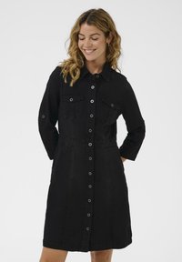 Vestido camisa de mezclilla negra con cierre de botones, dos bolsillos en el pecho, cuello y mangas de tres cuartos. Textura suave y corte ajustado.