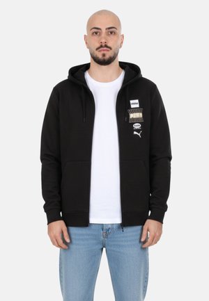 Hombre con barba que lleva una sudadera negra con cremallera de Puma sobre una camiseta blanca y vaqueros azul claro, de pie contra un fondo liso y claro.