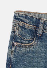 Jean en denim bleu délavé avec une petite poche avant, des coutures en cuivre et un accent métallique de marque sur la poche.
