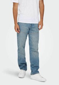 Blå denimjeans med en afslappet pasform, der har synlige stikninger, to forlommer og er parret med hvide sneakers og en plain hvid skjorte.
