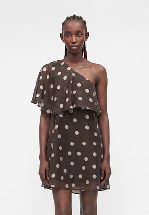 Μοντέλο που φοράει μαύρο φόρεμα με έναν ώμο και άσπρες πουά, με διαφανή στρωμάτωση βολάν στο στήθος και κοντό μήκος.