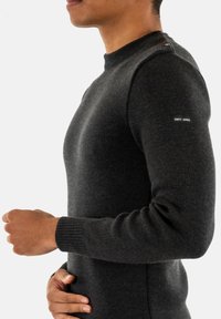 Maglione a maglia grigio scuro con scollatura rotonda, polsini a coste e dettagli testurizzati. Presenta bottoni decorativi sulla spalla. Etichetta del marchio visibile.