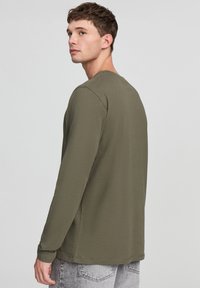 T-shirt verde oliva a maniche lunghe realizzato in tessuto morbido, con scollatura rotonda e vestibilità comoda, abbinato a jeans grigi chiari.