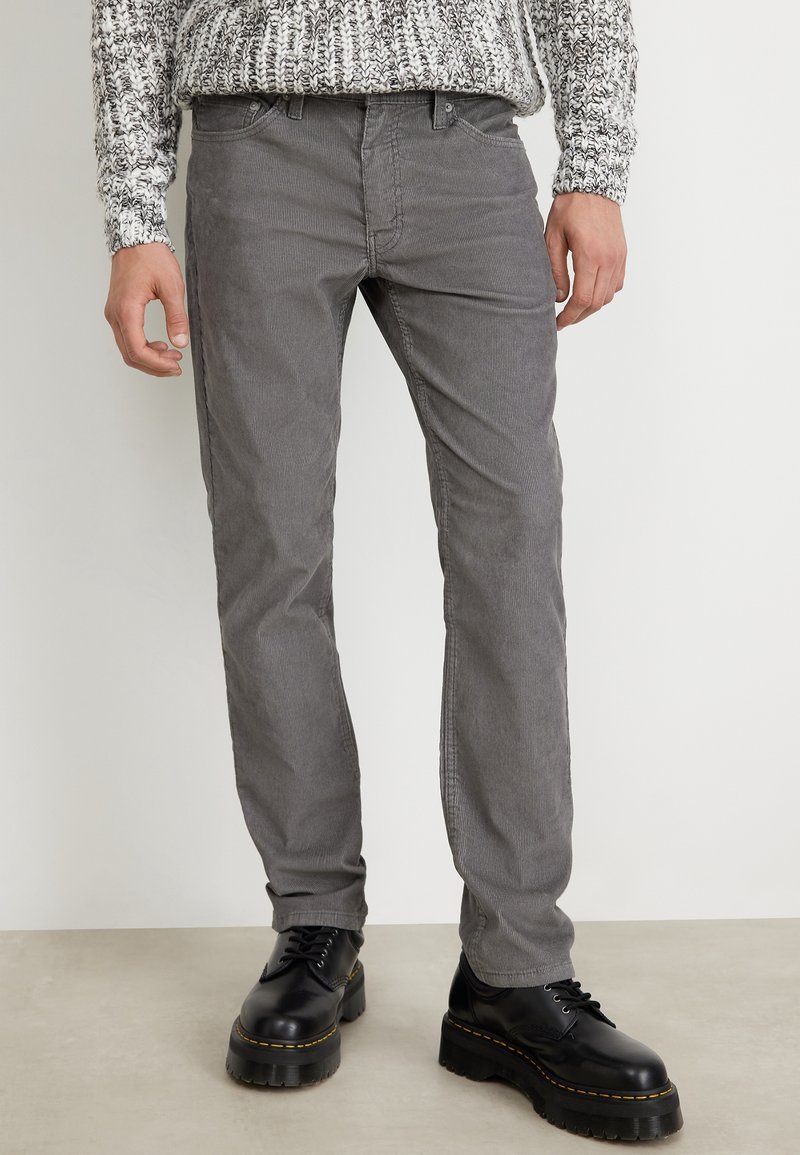 Homme portant un pantalon en velours côtelé gris, des chaussures noires épaisses à lacets et un pull en maille gris et blanc, debout à l’intérieur sur un sol clair.