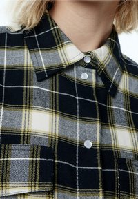 Karierte Hemd mit schwarz-weißen und gelben Mustern, mit Kragen, zwei Taschen und Kunststoffknöpfen zum Schließen. Textil mit Struktur.