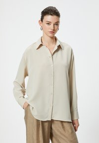 Camicia beige a maniche lunghe con chiusura a bottoni, colletto e texture a righe verticali; abbinata a pantaloni larghi di color marrone chiaro.
