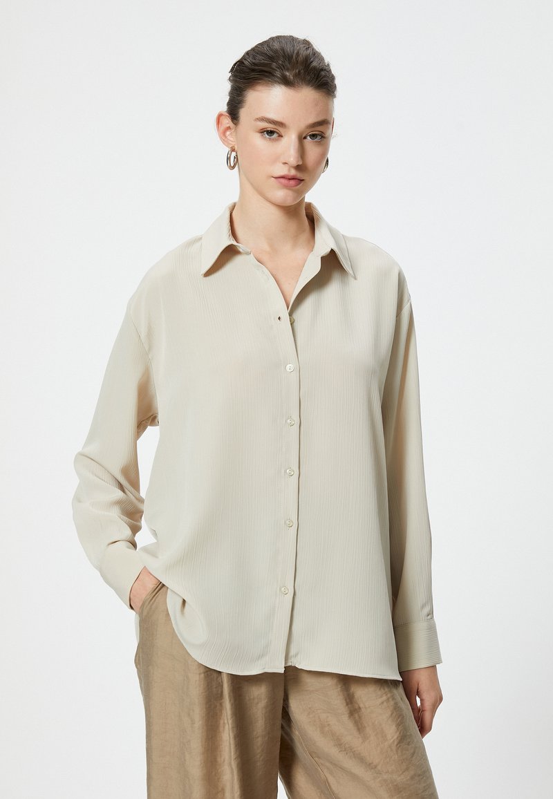 Camicia beige a maniche lunghe con chiusura a bottoni, colletto e texture a righe verticali; abbinata a pantaloni larghi di color marrone chiaro.