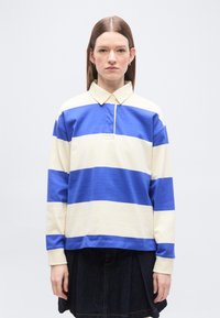 Femme aux cheveux bruns raides portant un maillot de rugby à larges rayures bleues et blanches et une jupe sombre, debout devant un fond clair uni.