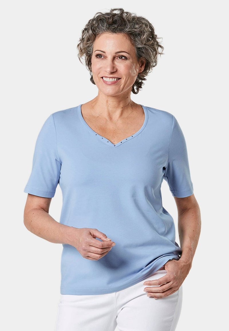 Lichtblauw T-shirt met korte mouwen en V-hals, versierd met studs bij de hals. Gemaakt van zachte stof met een losse pasvorm, gecombineerd met witte broeken.