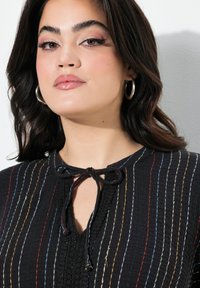 Blouse noire texturée avec des rayures verticales colorées, une fermeture à nouer au niveau du cou et des détails en dentelle délicate le long de la patte de boutonnage.