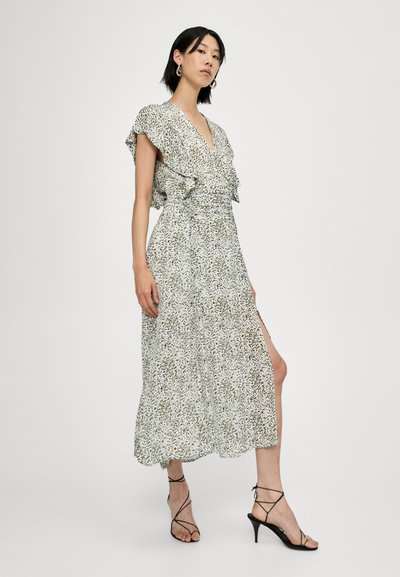 Robe maxi florale en vert clair avec des manches volantes, une taille cintrée et une fente sur le côté. Portée avec des escarpins à brides noirs.