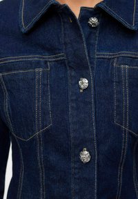 Tumesinine denimpusa, klassikalise krae, hõbedaste nööpide detailide ja kahe kõhupocketiga, millel on silmatorkav õmblus.
