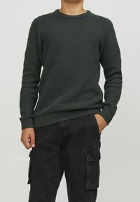 Man som bär en mörkgrön strukturerad stickad tröja och svarta cargobyxor, står med en hand i fickan mot en ljus bakgrund.