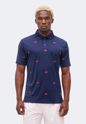 Modèle masculin aux cheveux blonds courts portant un polo bleu marine avec de petits motifs de chaussures rouges et un short de couleur claire, face à l'avant.