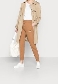 Beige trenchcoat, vit t-shirt, bruna sweats-byxor med logotyp och vita sneakers. Trenchcoaten har knappar och ett midjebälte.