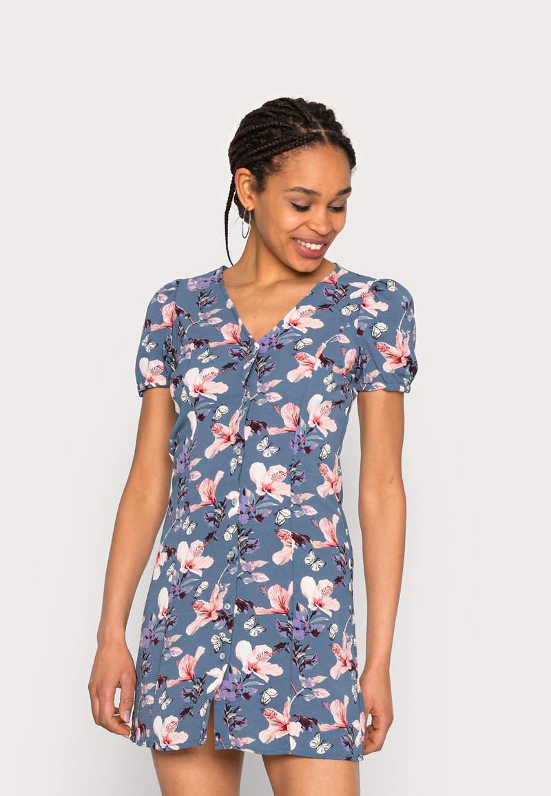Vestido de mangas curtas em tecido azul com um padrão floral, apresentando flores rosa e brancas e borboletas, decote em V e detalhe de cintura ajustada.