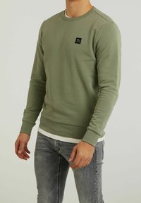 Homme portant un sweat-shirt à manches longues olive à col rond avec un petit patch logo noir et un jean gris délavé, debout devant un fond uni.