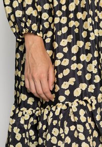 Schwarzes Kleid mit gelbem Blumenmuster, gerafften Ärmeln und Volant-Saum. Der Stoff hat eine glatte, glänzende Textur. Eine Hand, die den Stoff hält, ist sichtbar.