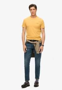 Gul bomuld T-shirt, tætsiddende design, med en strikket beige sweater bundet om taljen. Mørk vasket slim-fit jeans og sorte loafers.