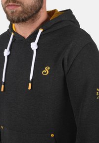 Sweat à capuche gris avec cordons blancs et accents jaunes. Dispose d'une poche avant et d'un logo brodé en jaune. Texture en mélange de coton.
