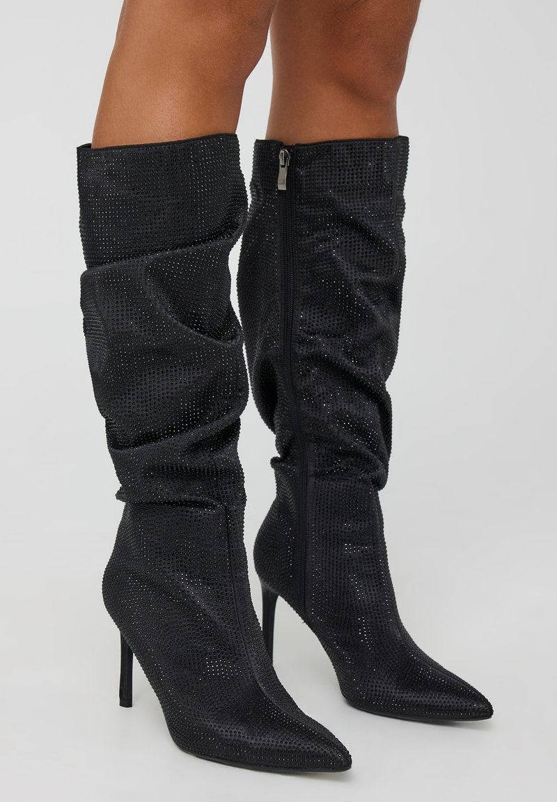 Stivali neri sopra il ginocchio con un design sgualcito, realizzati in tessuto scintillante con strass, punta affusolata e tacco a stiletto. Chiusura con zip laterale.