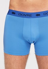Blå boxershorts med en glat tekstur, udstyret med et kontrasterende blåt elastikbånd med "DOVRE" og et lille logo. Strækbart og tætsiddende design.